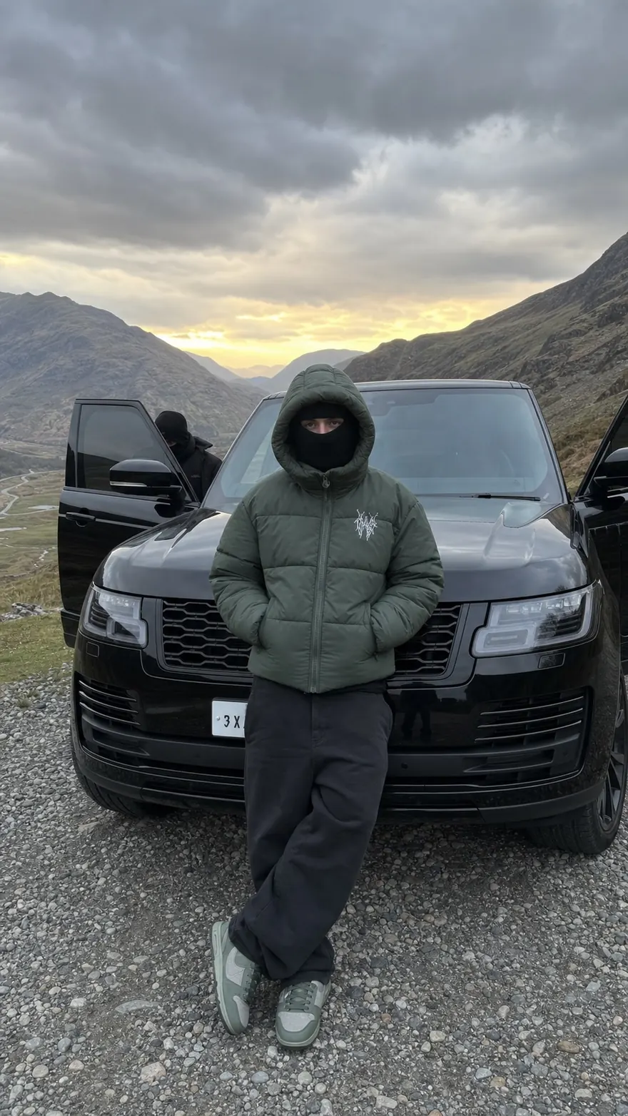 Blacked-Out Range Rover: Masked in a green puffer against a black Range Rover at dusk